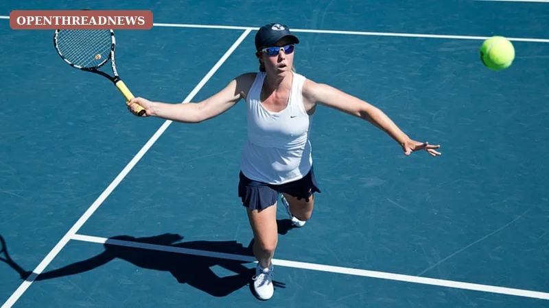 A college tennis player mid-serve in a navy and white kit on an outdoor court, representing Mayci Neeley's years playing tennis at BYU